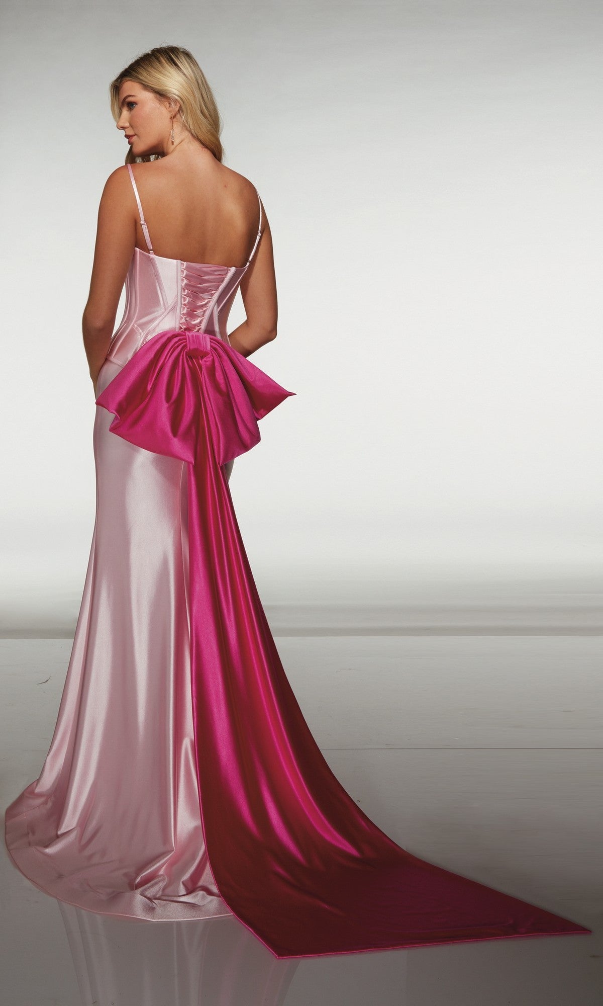 A woman wearing the Alyce 61983 long prom dress, featuring a sleeveless light pink satin design with a fitted corset bodice, large pink bow, and long train, stands with her back to the camera.